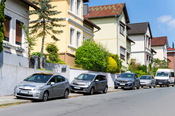 A row of cars parked along a sloped street lined with vintage, tall single-family homes. The houses are painted in friendly colors and are surrounded by green bushes and trees. A sunny day highlights the relaxed suburban atmosphere. This image is ideal for topics such as urban living, neighborhood life, and residential real estate.